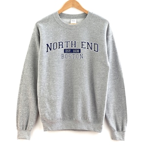 May include: A grey sweatshirt featuring the text "NORTH END" in navy blue, with "EST. 1630" and "BOSTON" underneath. The sweatshirt is hanging on a wooden hanger against a white backdrop.