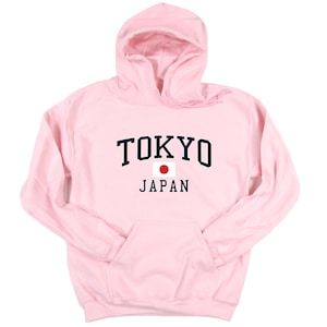 Peut inclure: Un sweat &agrave; capuche rose clair avec une poche avant et une capuche. Le mot "TOKYO" est imprim&eacute; en lettres majuscules bleu marine au-dessus du mot "JAPAN" en noir, avec un drapeau japonais entre les deux. Le sweat &agrave; capuche est en mati&egrave;re douce.