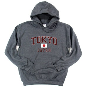 Peut inclure: Un sweat &agrave; capuche gris anthracite avec le mot "TOKYO" en lettres majuscules bordeaux au-dessus du mot "JAPAN". Le drapeau japonais est centr&eacute; entre les mots. Le sweat-shirt a une poche avant et une capuche &agrave; cordon.