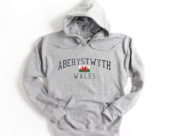 Puede incluir: Una sudadera con capucha gris con el texto "ABERYSTWYTH WALES" en letras azul marino. Un dragón rojo con fondo blanco y verde está centrado sobre el texto.