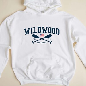 Pu&ograve; includere: Felpa con cappuccio bianca con una grafica blu navy del testo "WILDWOOD" con remi incrociati e una piccola bandiera americana. Il testo "19" e "12" sono su entrambi i lati dei remi. Il testo "NEW JERSEY" &egrave; sotto i remi.