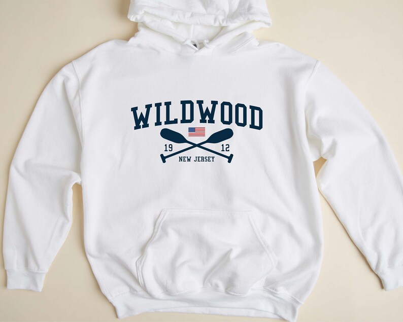 Pu&ograve; includere: Felpa con cappuccio bianca con una grafica blu navy del testo "WILDWOOD" con remi incrociati e una piccola bandiera americana. Il testo "19" e "12" sono su entrambi i lati dei remi. Il testo "NEW JERSEY" &egrave; sotto i remi.