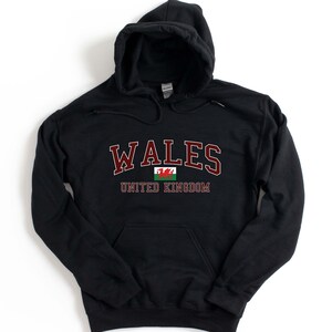 Puede incluir: Sudadera con capucha negra con el texto "WALES UNITED KINGDOM" en letras granate y un gr&aacute;fico de la bandera del drag&oacute;n rojo.