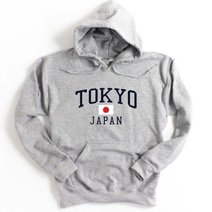 Peut inclure: Un sweat-shirt &agrave; capuche gris clair avec une poche avant et un cordon de serrage. Le mot "TOKYO" est imprim&eacute; en bleu marine en arc au-dessus de "JAPAN" en noir, avec un drapeau japonais entre les deux.