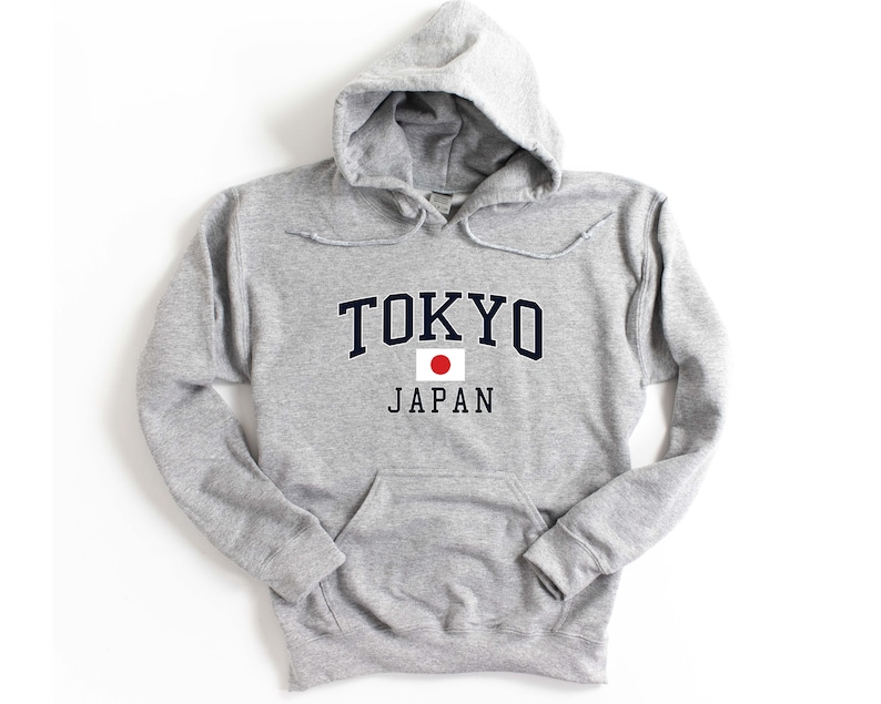 Peut inclure: Un sweat-shirt &agrave; capuche gris clair avec une poche avant et un cordon de serrage. Le mot "TOKYO" est imprim&eacute; en bleu marine en arc au-dessus de "JAPAN" en noir, avec un drapeau japonais entre les deux.