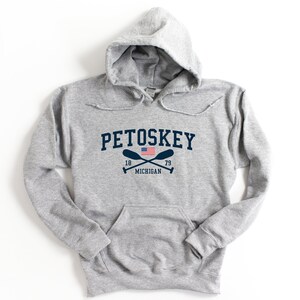 May include: A gray hooded sweatshirt with the text "Petoskey" in navy blue lettering, a small American flag, and two crossed oars with the text "18 79 MICHIGAN" underneath.