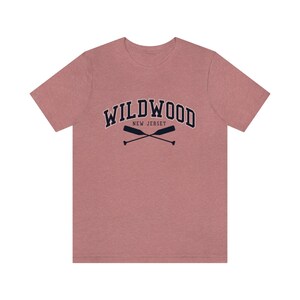 May include: A heather pink t-shirt with a navy blue graphic of the text "WILDWOOD NEW JERSEY" in an arched font with two crossed oars underneath.