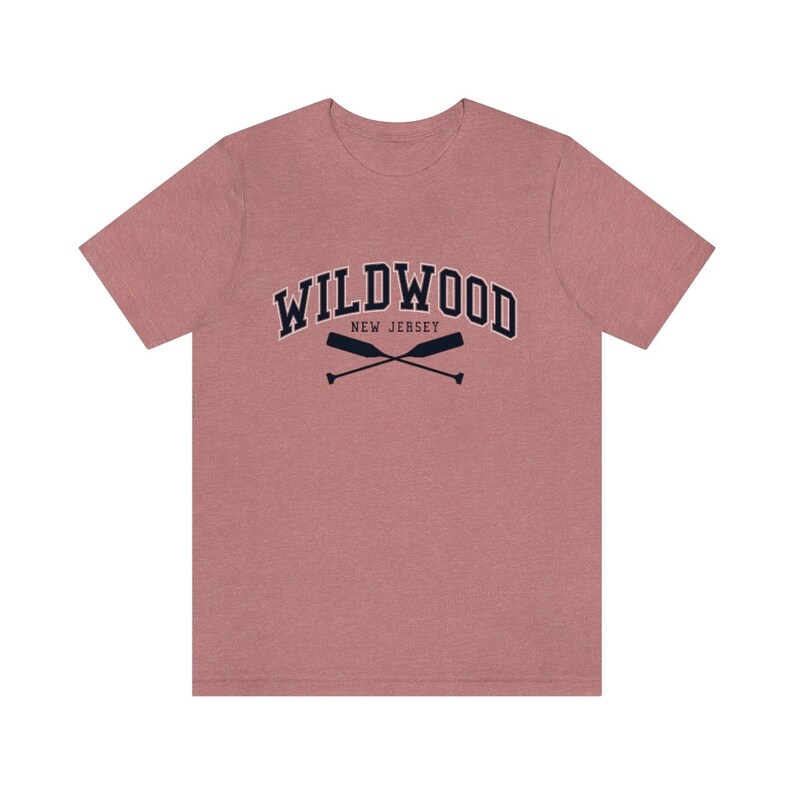 May include: A heather pink t-shirt with a navy blue graphic of the text "WILDWOOD NEW JERSEY" in an arched font with two crossed oars underneath.