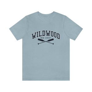 May include: A light blue t-shirt with a navy blue graphic of the text "WILDWOOD NEW JERSEY" in a collegiate font with two crossed oars underneath.