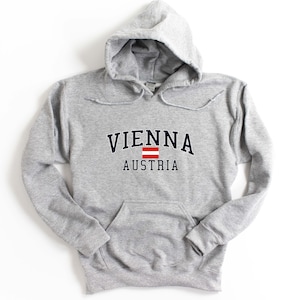 May include: A light gray hooded sweatshirt with a front pocket and drawstring. The word "VIENNA" is arched across the chest in navy blue letters, with "AUSTRIA" below. A red and white Austrian flag is between the words.