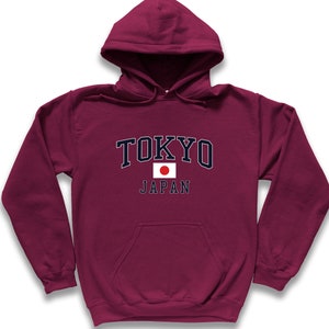 Peut inclure: Un sweat &agrave; capuche bordeaux avec le mot "TOKYO" en lettres blanches avec un contour bleu marine. En dessous du mot "TOKYO" se trouve le mot "JAPAN" et un petit drapeau japonais. Le sweat-shirt a une poche avant.