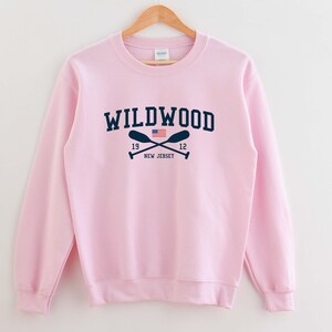 May include: Pink sweatshirt with a navy blue graphic of the text "WILDWOOD" with crossed oars and a small American flag. The text "19" and "12" are on either side of the oars. The text "NEW JERSEY" is below the oars.