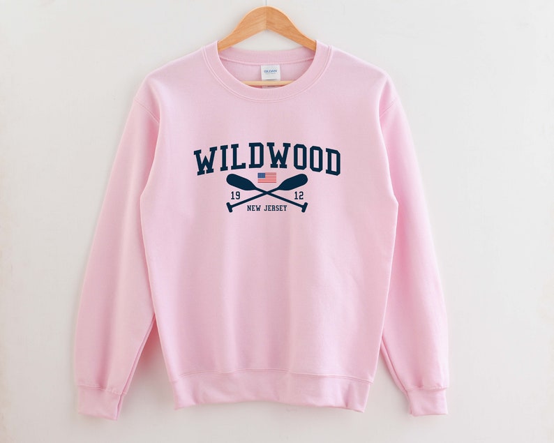 May include: Pink sweatshirt with a navy blue graphic of the text "WILDWOOD" with crossed oars and a small American flag. The text "19" and "12" are on either side of the oars. The text "NEW JERSEY" is below the oars.