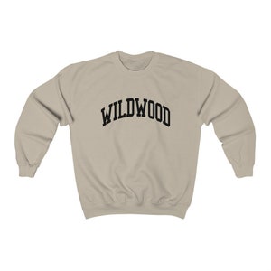 May include: A beige sweatshirt with the word "WILDWOOD" arched across the chest in bold, black letters. The sweatshirt has a crew neck and long sleeves, suitable for casual wear.