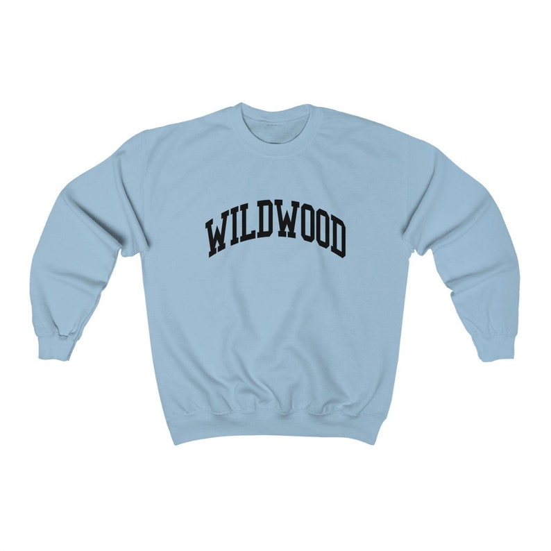 May include: A light blue sweatshirt with the word "WILDWOOD" printed in black, arched letters. The sweatshirt has long sleeves and a crew neck. The fabric appears to be a soft, comfortable material.