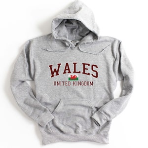 Puede incluir: Sudadera gris con capucha con la palabra "WALES" en letras marrones y un peque&ntilde;o gr&aacute;fico de la bandera galesa debajo. El texto "UNITED KINGDOM" est&aacute; impreso en marr&oacute;n debajo de la bandera.