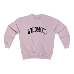 May include: A light pink sweatshirt with the word "WILDWOOD" arched across the chest in black block letters. The sweatshirt has long sleeves and a crew neck. The fabric appears to be a soft, comfortable material.