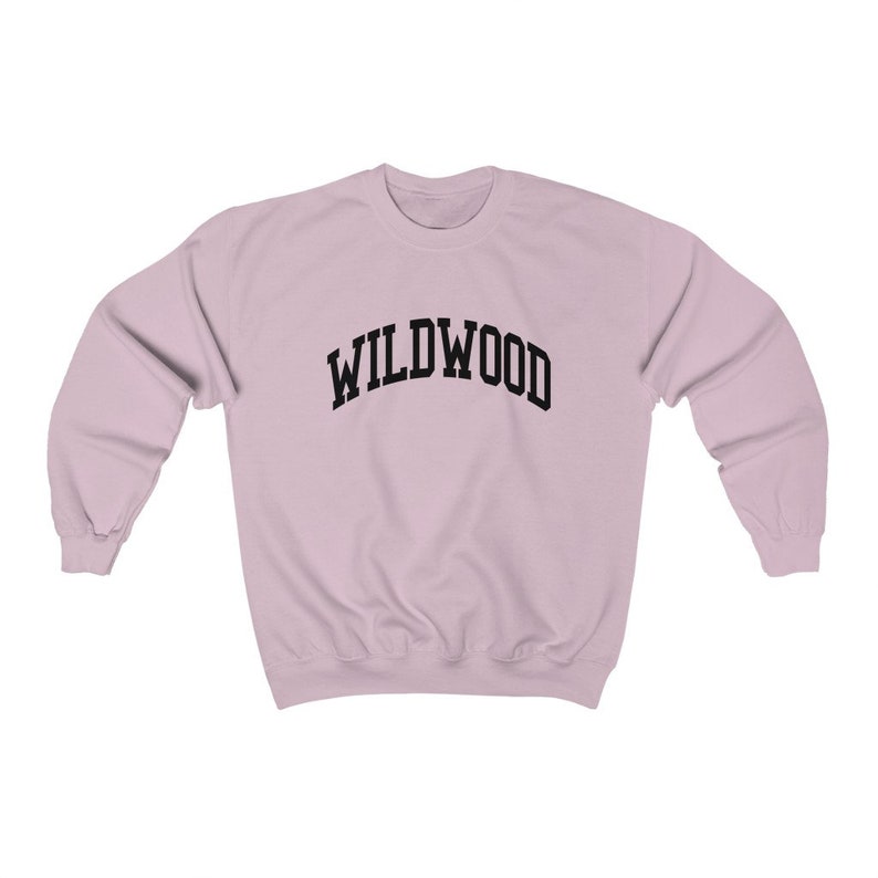 May include: A light pink sweatshirt with the word "WILDWOOD" arched across the chest in black block letters. The sweatshirt has long sleeves and a crew neck. The fabric appears to be a soft, comfortable material.