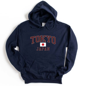 Peut inclure: Un sweat-shirt &agrave; capuche bleu marine avec le mot "TOKYO" en lettres majuscules bordeaux avec un contour blanc. En dessous, un petit drapeau japonais et le mot "JAPAN" en blanc.
