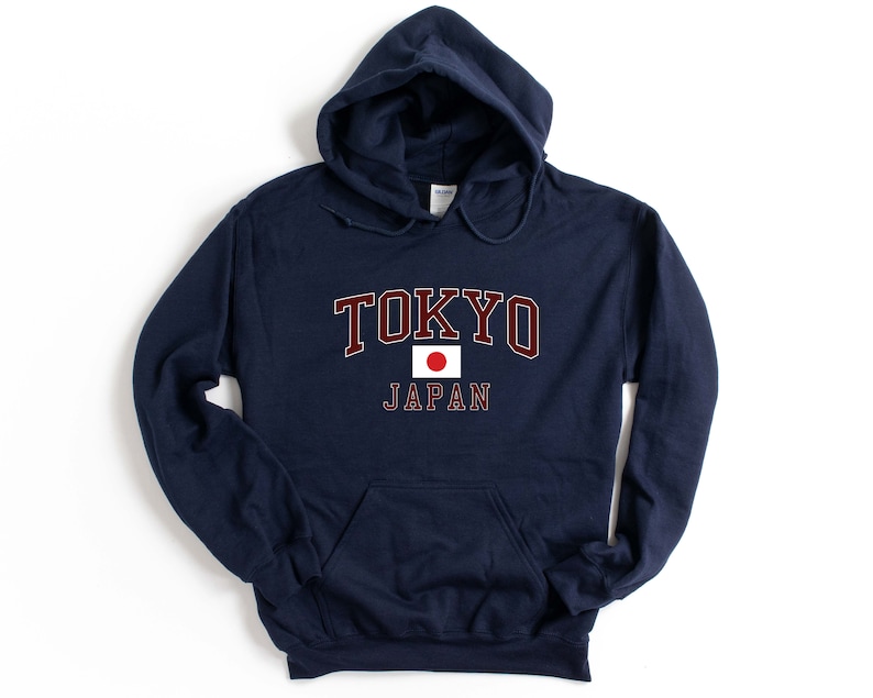 Peut inclure: Un sweat-shirt &agrave; capuche bleu marine avec le mot "TOKYO" en lettres majuscules bordeaux avec un contour blanc. En dessous, un petit drapeau japonais et le mot "JAPAN" en blanc.