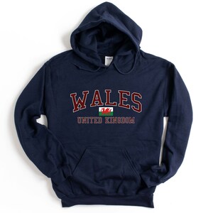 Puede incluir: Una sudadera con capucha azul marino con la palabra "WALES" en letras granate y la bandera galesa debajo. El texto "UNITED KINGDOM" est&aacute; impreso debajo de la bandera.