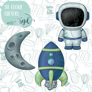 Pode incluir: Um conjunto de três cortadores de biscoitos em forma de astronauta, foguete e lua crescente. O astronauta está vestindo um traje espacial branco com uma viseira azul. O foguete é azul e amarelo com uma chama branca. A lua é cinza com crateras.