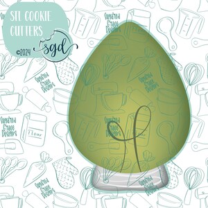 Puede incluir: Un cortador de galletas verde en forma de árbol de Navidad con una base blanca. El cortador está sobre un fondo blanco con un patrón de utensilios de cocina y el texto "Sugared Grace Designs".