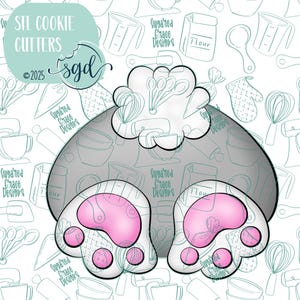 May include: A gray cookie cutter in the shape of a cat's rear end with two pink paw prints. The cutter is surrounded by a white background with a pattern of kitchen tools and the text "Suggared Grace Designs".