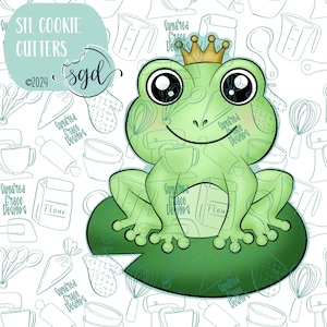 May include: A green frog wearing a gold crown sits on a green lily pad. The frog has large, black eyes and a wide smile. The image is surrounded by a pattern of kitchen tools and baking supplies.