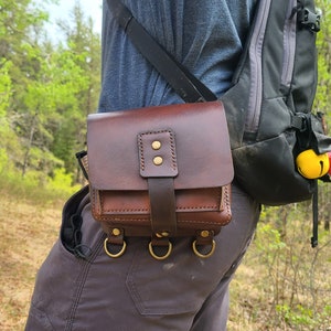 May include: A brown leather belt bag with a strap and three metal rings. The bag has a flap with two brass buttons and is worn on a person's waist.