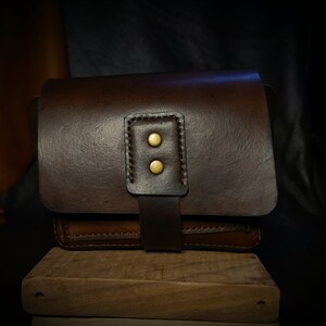 May include: A brown leather bag with a flap closure and two brass button accents. The bag is sitting on a wooden surface.