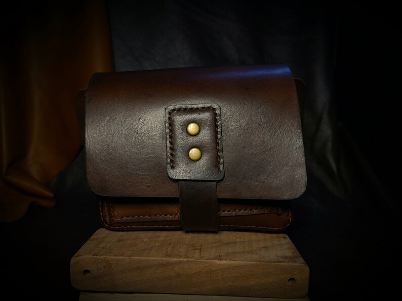 May include: A brown leather bag with a flap closure and two brass button accents. The bag is sitting on a wooden surface.