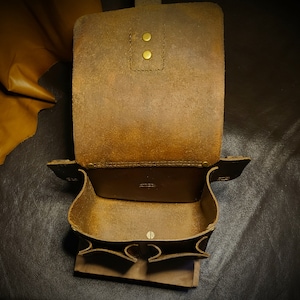 May include: A brown leather bag with two pockets and a flap closure. The bag is open and the inside is visible. The bag is made of a thick, durable leather and has a rustic look.