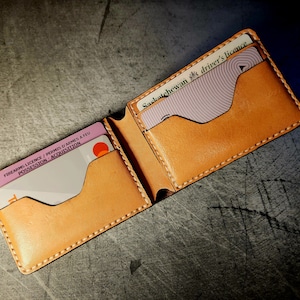 May include: A tan leather wallet with two card slots. The wallet is open and shows a white card with the text "FIREARMS LICENCE / PERMIS D'ARMES A FEU POSSESSION - ACQUISITION" and a pink card with the text "Saskatchewan driver's licence".