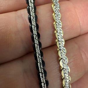以下が含まれることがあります： 2つのブレスレットのクローズアップ。1つは黒でシルバーカラーのチェーン、もう1つはゴールドカラーでシルバーカラーのチェーンです。どちらも、小さく接続された円形のリンクの繰り返しパターンを持っています。