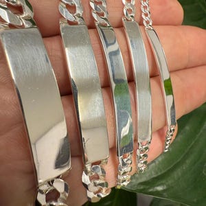 Puede incluir: Cinco pulseras de cadena curb plateadas con un dije rectangular en forma de placa. Las placas están en blanco y listas para ser grabadas.