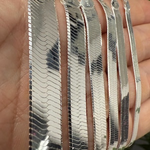 Puede incluir: Varias cadenas de plata brillantes con diseño de espiga. Los collares presentan un diseño plano y tejido. Las cadenas se sostienen en una mano y el metal tiene una superficie reflectante. Las cadenas están marcadas con "925 ITALY".