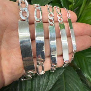 May include: Five silver ID bracelets with different chain styles. The bracelets have a flat, rectangular tag that can be engraved.