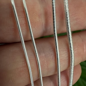 May include: Close-up of four silver snake chains. The chains vary slightly in thickness, with a smooth, reflective surface. The chains are displayed against a blurred background, highlighting their intricate design and metallic sheen. Ideal for jewelry making.