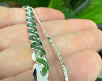 May include: Two silver chains, one with a wavy pattern and the other with a delicate, twisted design. The chains are held in a hand against a green background.