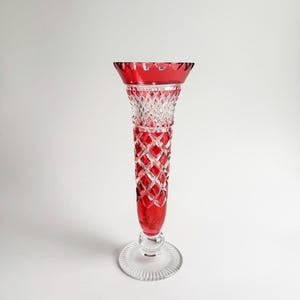 May include: A ruby red crystal vase with a diamond pattern on the stem and a scalloped rim. The vase has a clear, circular base and stands approximately 8 inches tall.