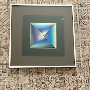May include: Framed abstract art print featuring a geometric design with concentric squares in shades of blue, purple, and white. The artwork is set against a dark gray background and framed in a white border. The piece is a modern art piece.