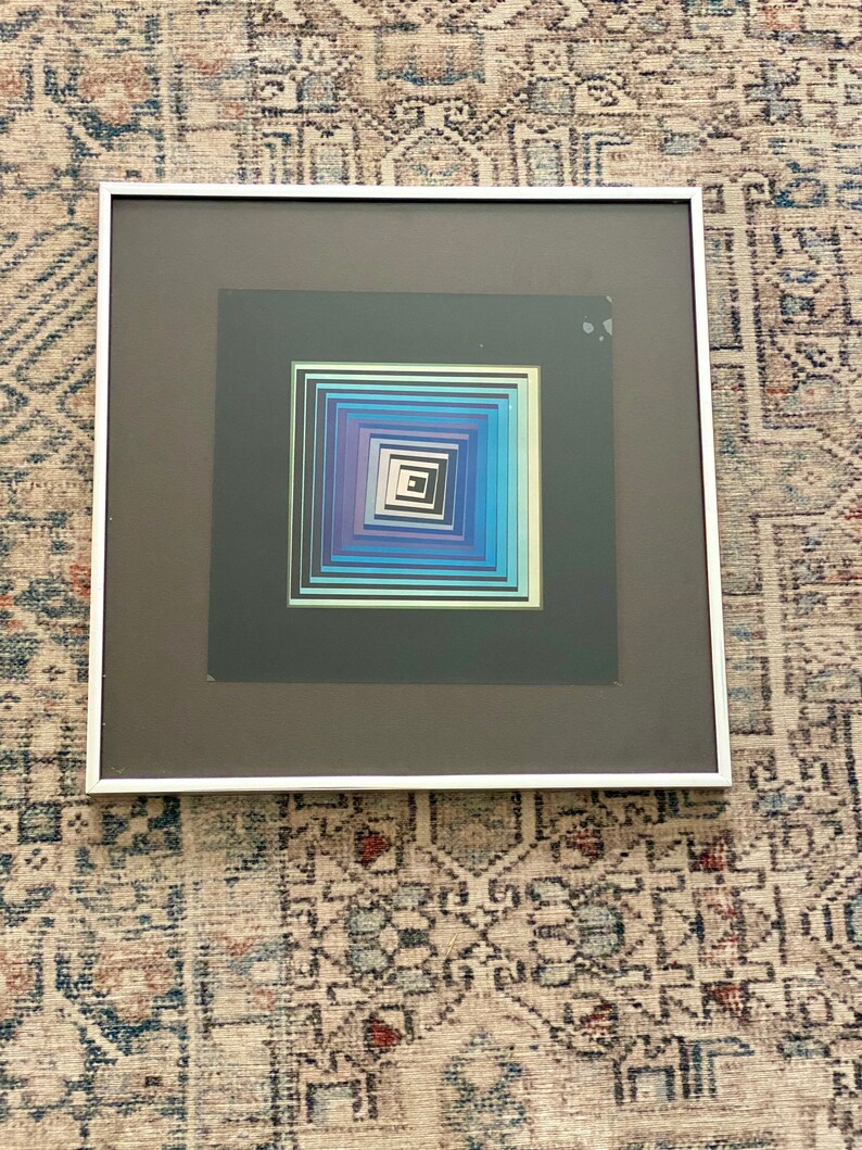 May include: Framed abstract art print featuring a geometric design with concentric squares in shades of blue, purple, and white. The artwork is set against a dark gray background and framed in a white border. The piece is a modern art piece.