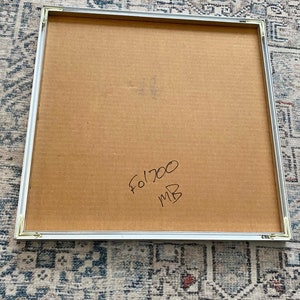 May include: A square framed artwork with a cardboard backing. The frame is silver. Handwritten in black ink on the cardboard is "Fo1700 MB". The artwork is resting on a patterned rug.