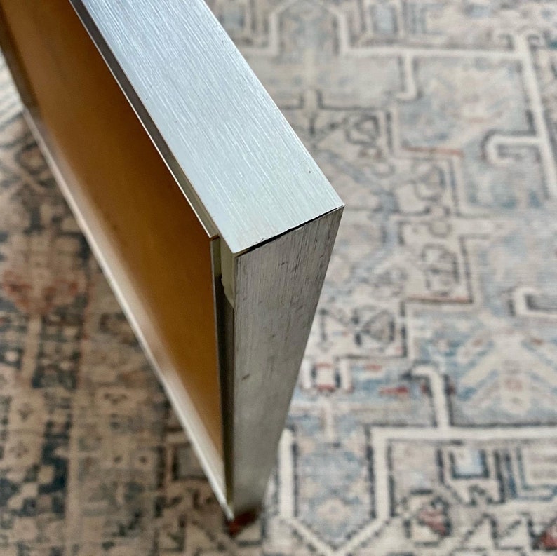 May include: Close-up of a silver metal picture frame with a light brown interior. The frame has a brushed metal finish and a clean, modern design. The corner of the frame is visible, showing the precise joinery.