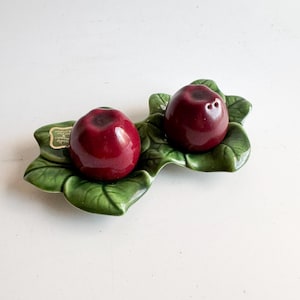 May include: A ceramic salt and pepper set featuring two glossy, deep red apple-shaped shakers. The shakers rest on a green leaf-shaped base. The set is designed to resemble a pair of apples on a bed of leaves.
