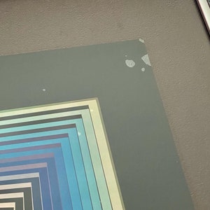 May include: Framed abstract art print featuring a series of nested squares in shades of blue, teal, and gray. The artwork is encased in a white border and a gray frame, creating a modern aesthetic. The print is displayed against a gray wall.