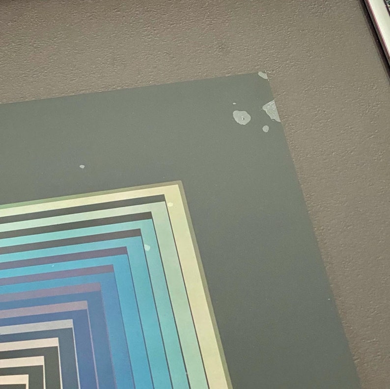 May include: Framed abstract art print featuring a series of nested squares in shades of blue, teal, and gray. The artwork is encased in a white border and a gray frame, creating a modern aesthetic. The print is displayed against a gray wall.