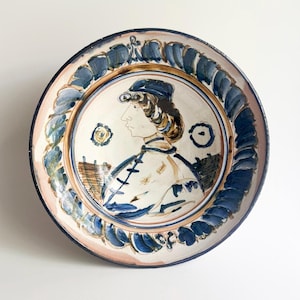 May include: A round ceramic plate featuring a hand-painted profile portrait of a person wearing a cap. The design uses blue, brown, and cream tones, with a decorative border of blue brushstrokes encircling the central figure and two small circles.