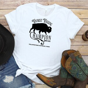 May include: White t-shirt with a black graphic design of a bison silhouette and the text "Tourist Tossing Champion Since 1872 Yellowstone National Park".